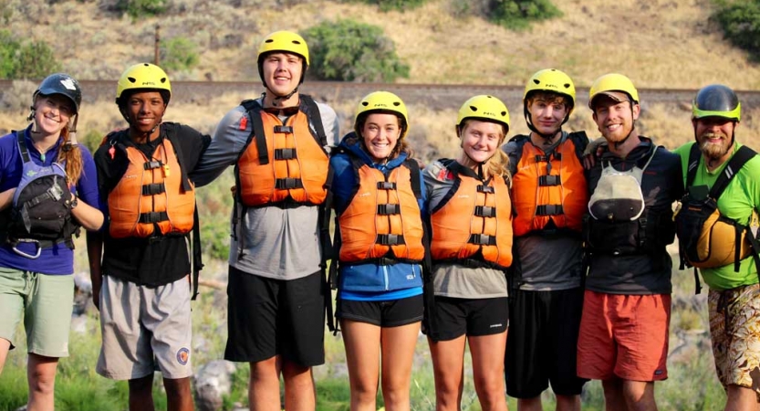 a group of students wearing helmets and life jackets stand and with their arms around each other and smile at the camera